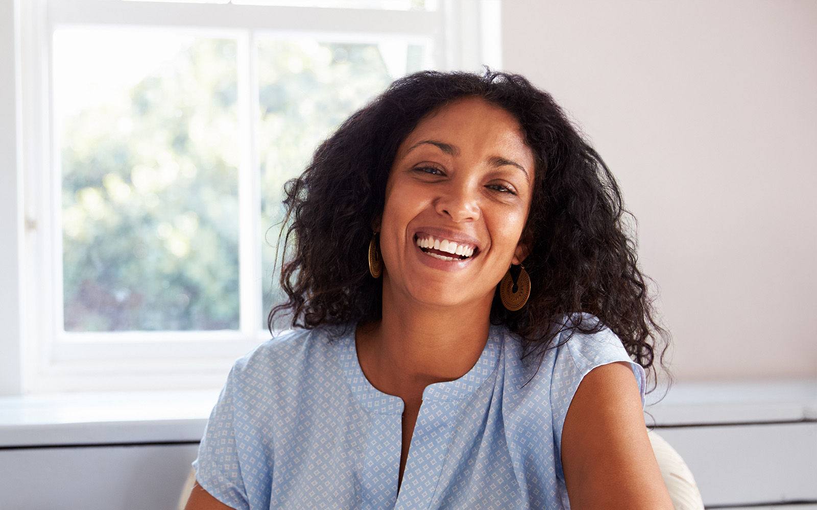 A woman with a radiant smile, wearing a light blue blouse, sits in front of a window.