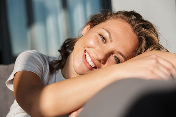 A woman with a radiant smile, resting her head on her arm while leaning against a couch.