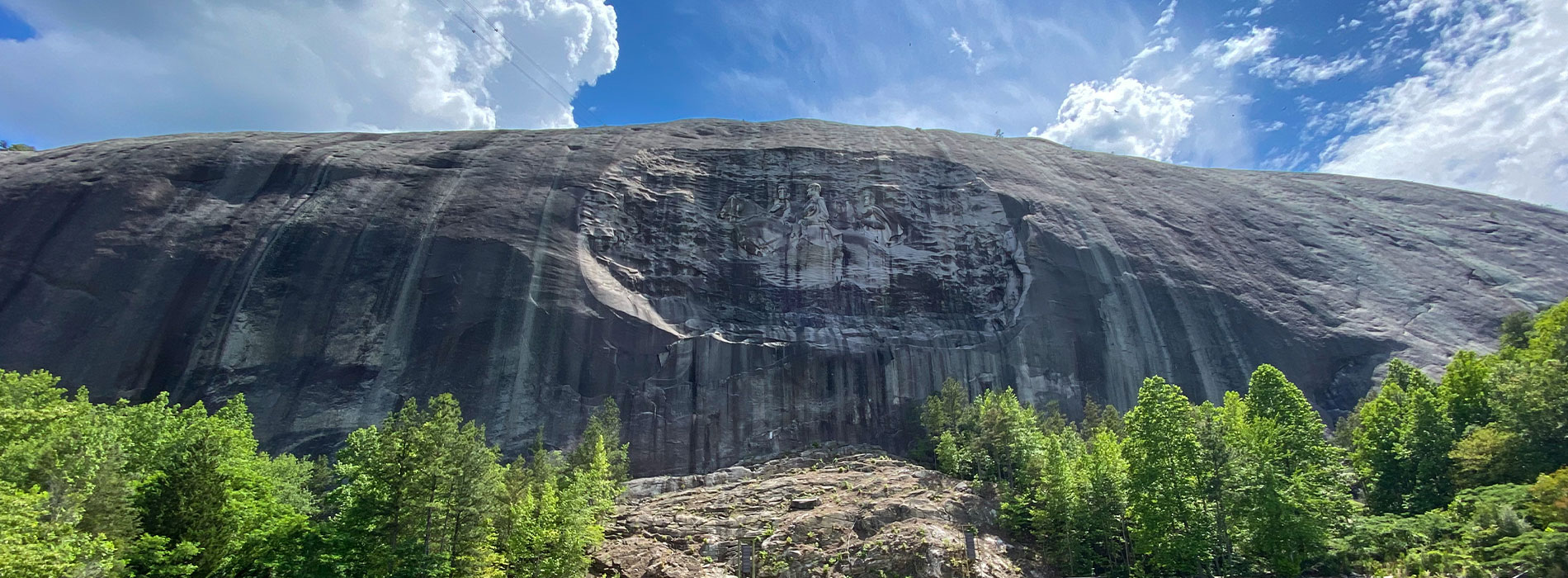 The image depicts a large, prominent rock formation with a flat top, situated in a natural setting with trees and a clear sky.