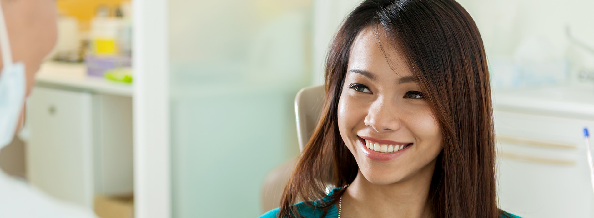 The image is a photograph of a woman with dark hair, smiling and looking at the camera while seated in a dental office chair. She appears to be having a dental examination or cleaning, as suggested by her relaxed posture and the presence of a dental professional partially visible behind her.