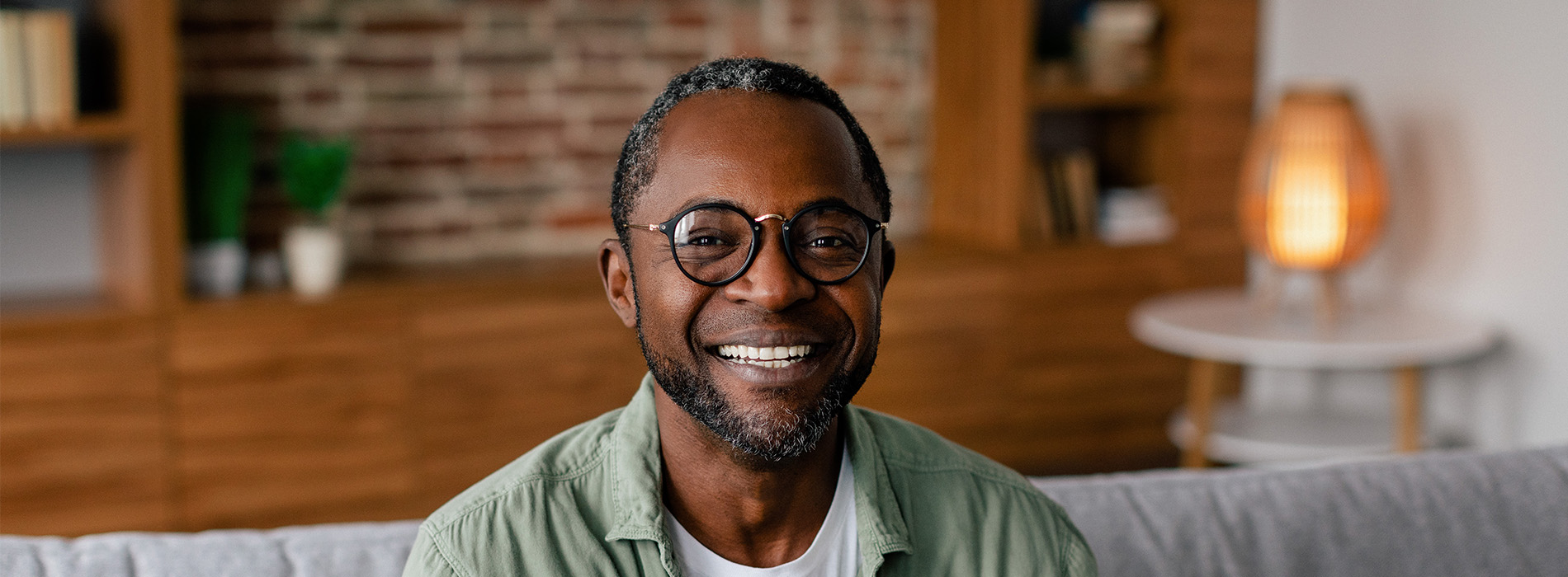 The image features a man sitting on a couch, smiling and looking directly at the camera. He has a beard and is wearing glasses. Behind him, there is a blurred background with a brick wall and some furniture.