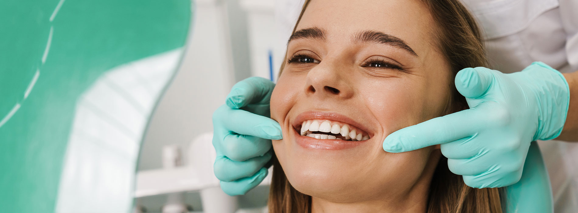 A woman is seated in a dental chair, smiling as a dentist adjusts her teeth with their hands.