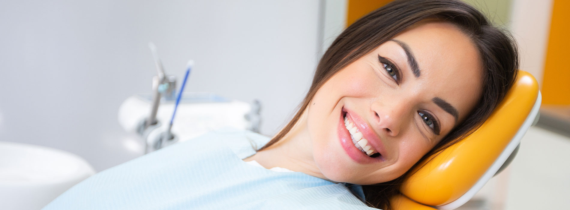 The image shows a smiling woman in a dental chair, with her head resting on a blue pillow, wearing a white face mask, and she appears to be receiving dental care.