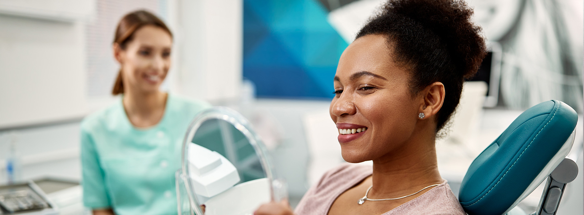 A woman in a dental office, smiling and seated in a dental chair, with a dental hygienist standing behind her.