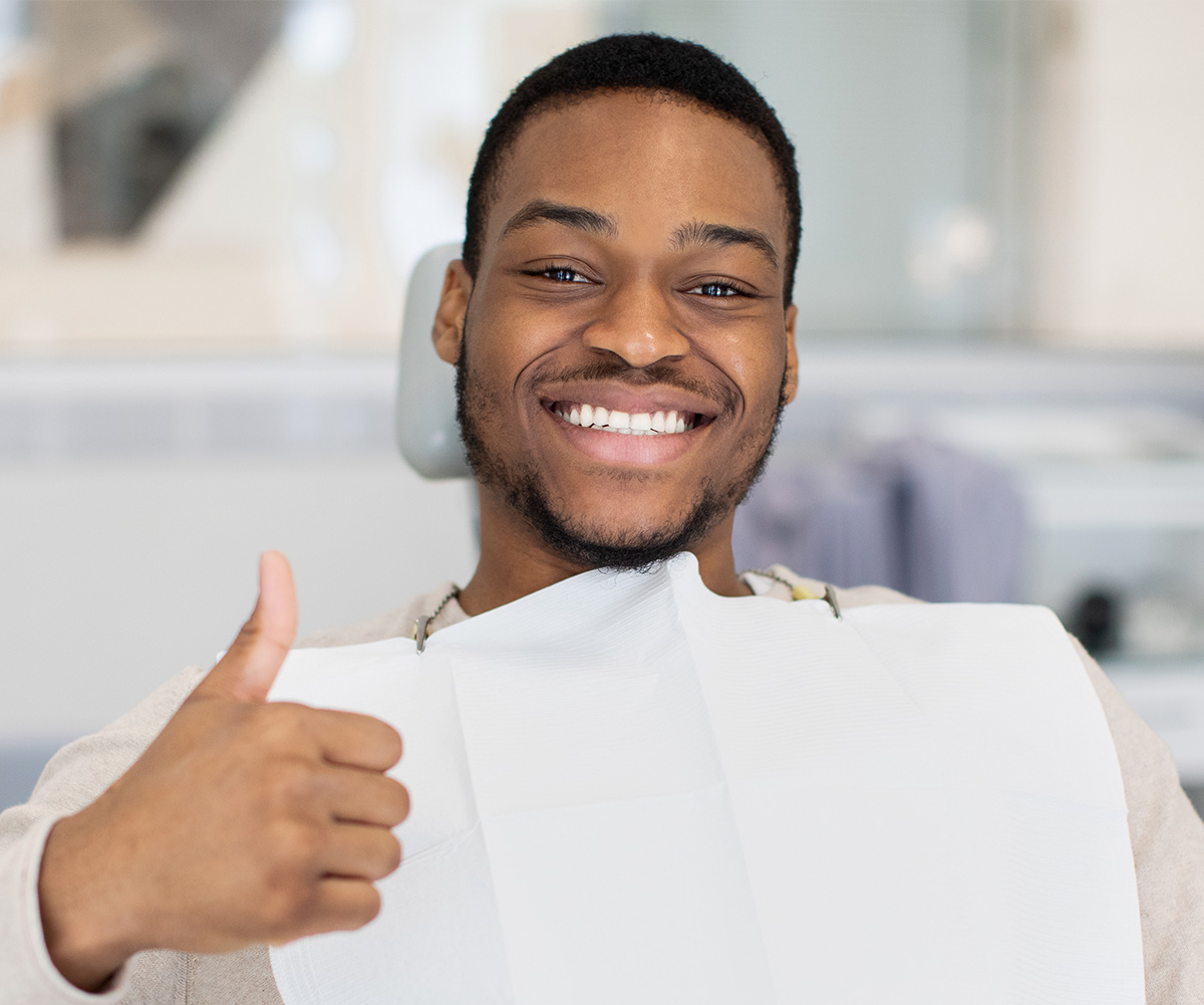 The image shows a person with a thumbs-up gesture, smiling and giving a thumbs-up sign in front of dental equipment.