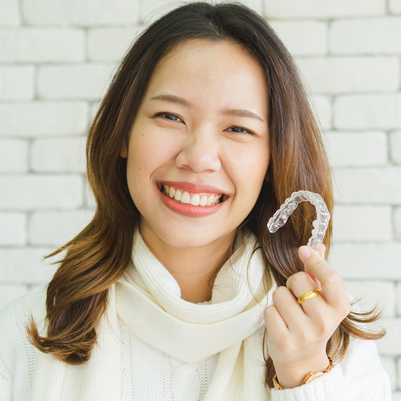 The image features a woman smiling at the camera while holding up a clear dental retainer with her left hand. She is indoors, wearing a white sweater and has dark hair.