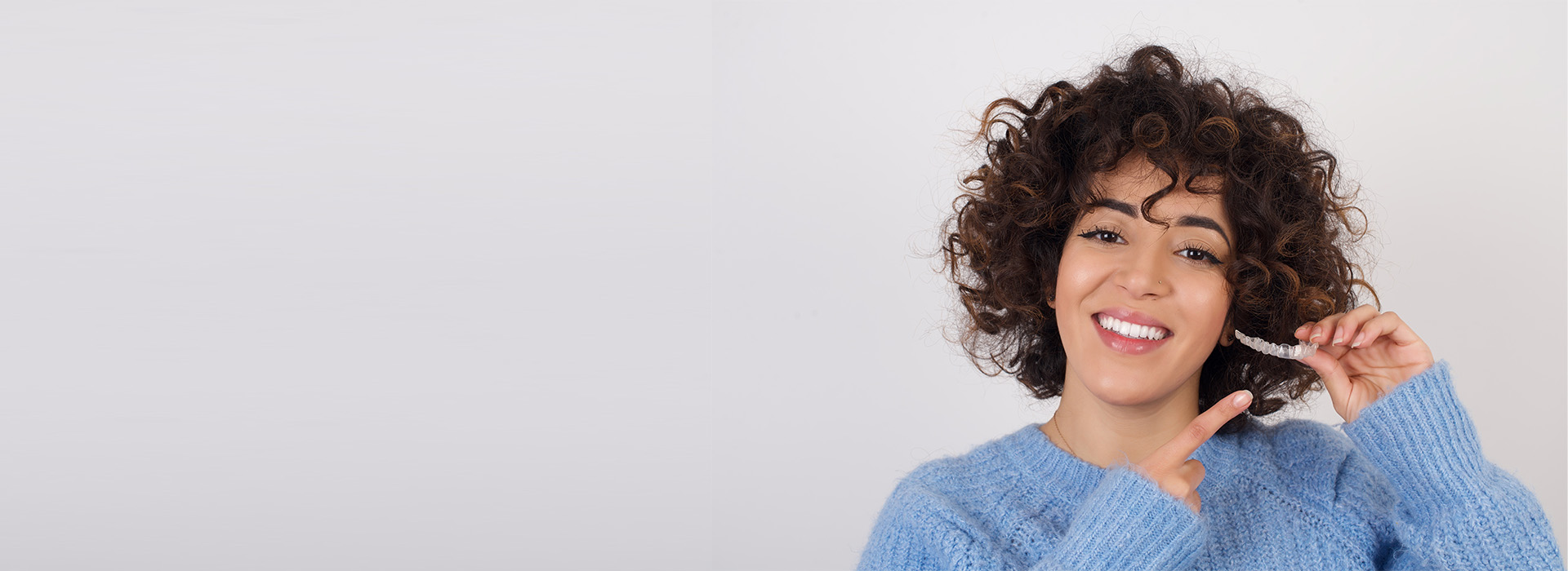 The image shows a smiling individual with curly hair, wearing a blue sweater and holding a phone to their ear. They are standing against a plain background.