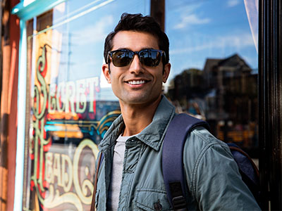 A man with sunglasses and a backpack stands in front of a store window, smiling.