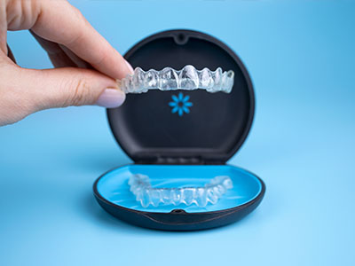 The image shows a person s hand holding an open clear case containing a set of transparent dental aligners, with the aligners partially inserted into a blue plastic mouthpiece that represents a human mouth.