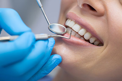 The image shows a dental professional performing teeth cleaning or oral care on a patient, with the patient wearing a blue surgical gown and having their mouth open, while the dental professional is using specialized tools and wearing gloves.