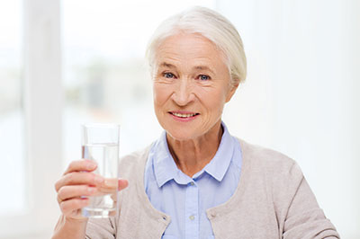 An elderly woman in a blue blazer, holding a glass of water and smiling at the camera.