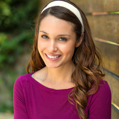 A woman smiling at the camera, wearing a purple top and headband, against a wooden fence background.