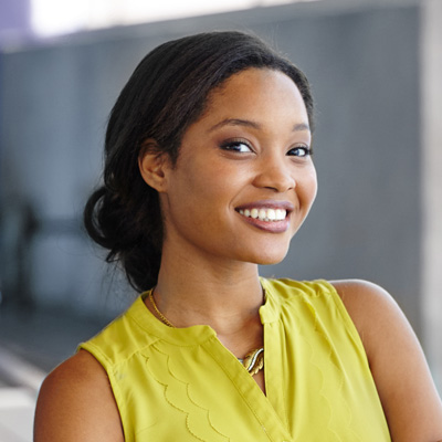 A young woman with a radiant smile, wearing a yellow top and standing against a backdrop of a building, poses for the camera.