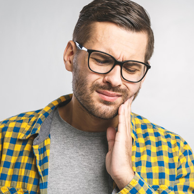 The image depicts a man wearing glasses, with his hand placed on his chin and his mouth open as if in mid-speech or thought. He has dark hair and is dressed in a yellow plaid shirt.