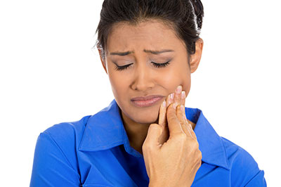 The image features a woman with dark hair, wearing a blue top, and making a face with her hand on her chin. She appears to be indoors and is looking downwards while holding her nose with her right hand.