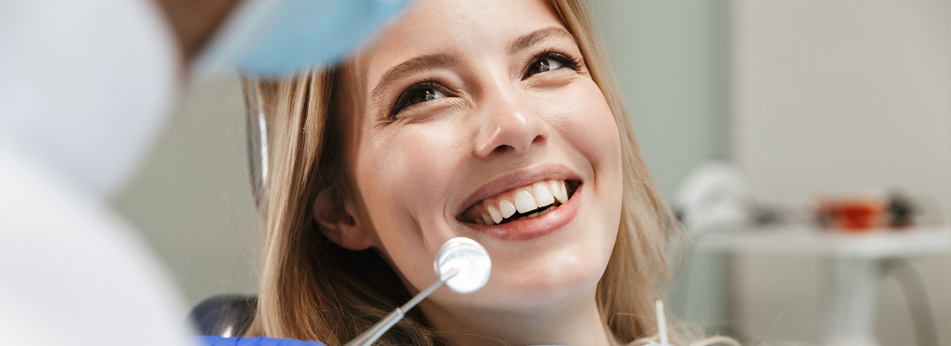 The image shows a smiling woman in a dental setting, receiving attention from a dental professional holding a mirror.