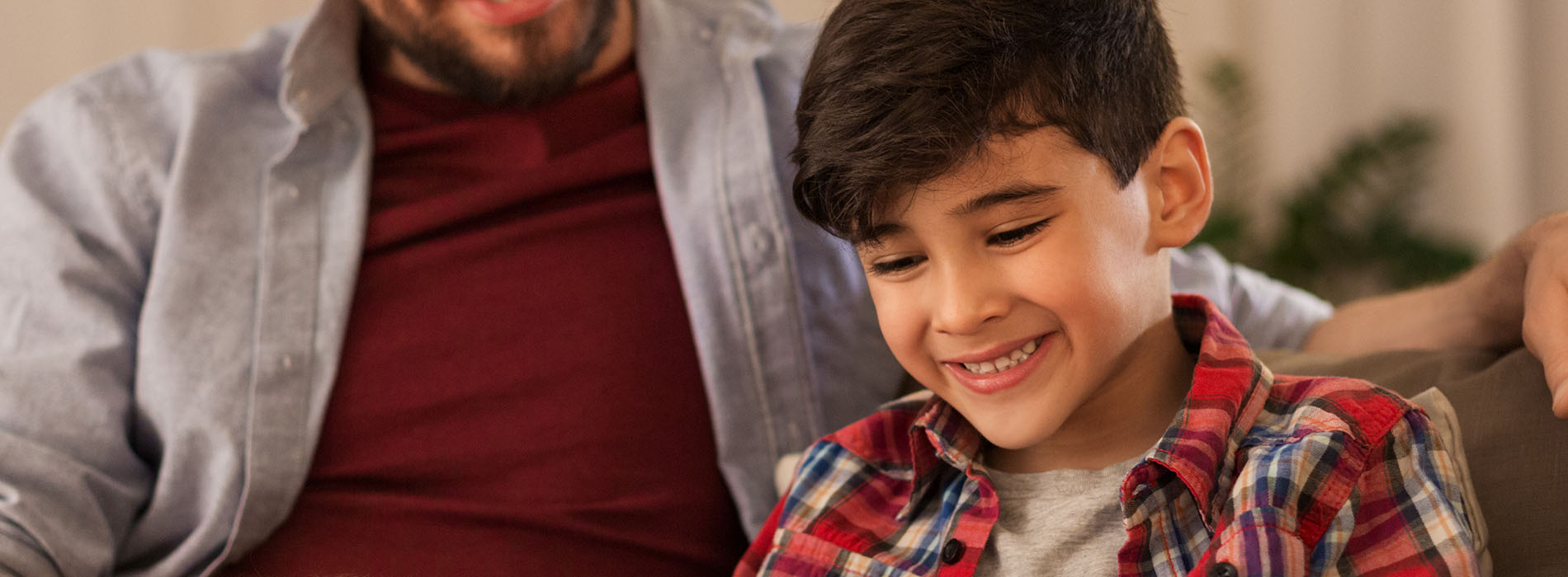 The image shows a man and a young boy sitting together on a couch, with the man smiling at the camera and the boy looking towards him. They appear to be in an indoor setting.