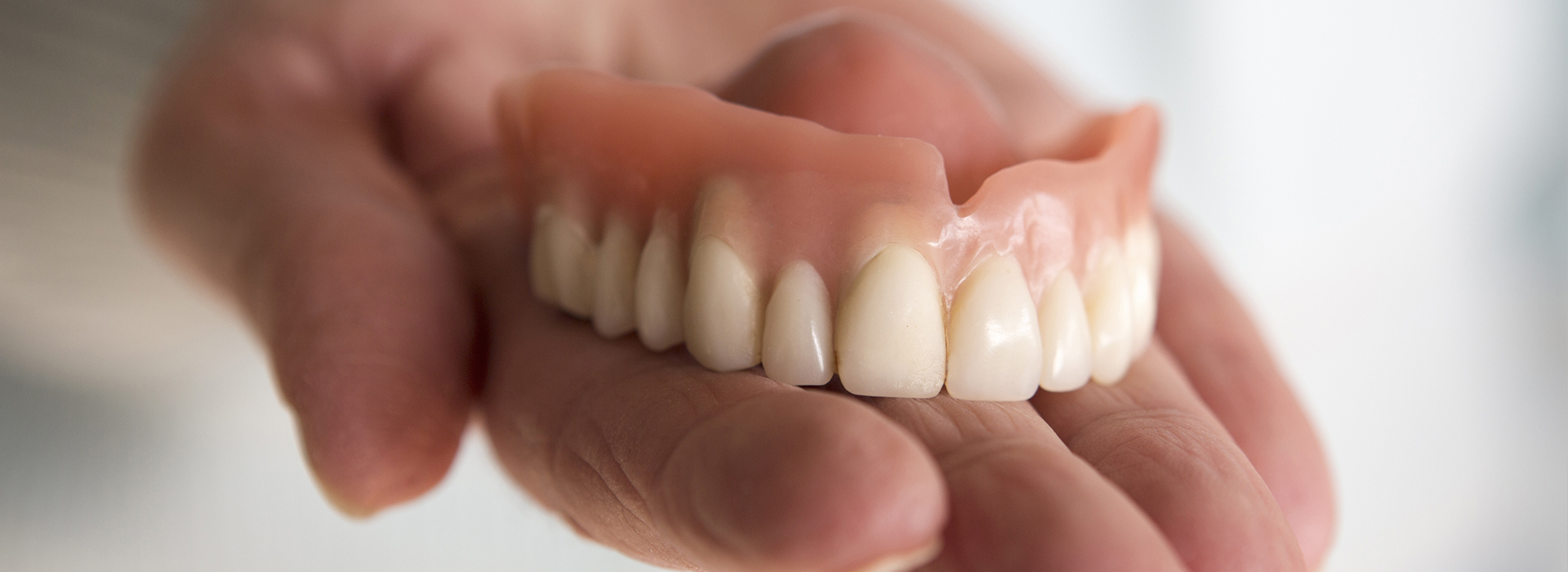 The image shows a person holding a set of dentures, which are white and appear to be for upper teeth.