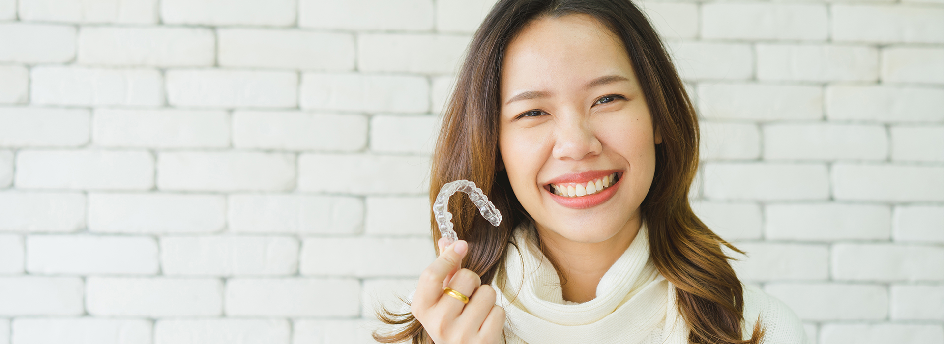 A smiling woman wearing a white top, holding a ring and standing in front of a brick wall.