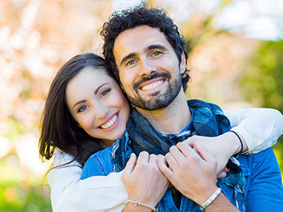 A man and a woman embracing in a park, both smiling.