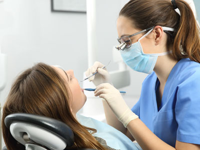 A dental hygienist in a blue gown and white gloves, wearing a face mask, is working on a patient s teeth at a dental office.