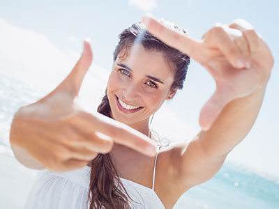 A young woman with a radiant smile is taking a selfie against a bright beach backdrop, holding her camera at arm s length.