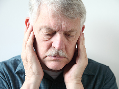 The image shows an older man with a mustache, wearing glasses and a gray shirt, holding his hand to his ear. He appears to be in pain or discomfort.